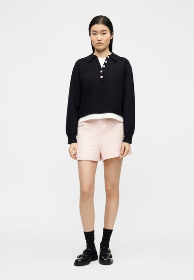 Γυναίκα που φοράει μαύρο μακρυμάνικο πουλόβερ με κουμπιά πάνω από λευκό τοπ, ροζ υφασμάτινη βερμούδα, μαύρες κάλτσες και μαύρα παπούτσια με αγκράφες, στέκεται όρθια.