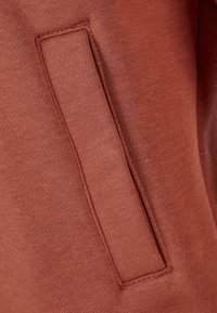 Tkanina rjave barve z gladko teksturo vsebuje detajl žepa z všitim šivom, ki poudarja funkcionalno zasnovo oblačila.