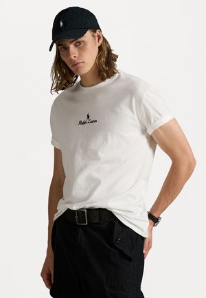 Witte katoenen T-shirt met opgerolde mouwen, voorzien van een zwart geborduurd logo, gecombineerd met zwarte cargoshorts en een zwarte pet.