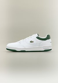 White leather sneaker with green accents, lace-up design, round toe, and textured sole. Features the Lacoste logo on the side and heel.