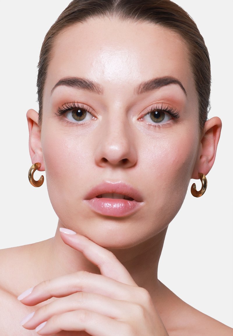 Orecchini ad anello a spirale color oro indossati da un modello con pelle liscia e trucco neutro, che mettono in evidenza i suoi tratti su uno sfondo semplice.