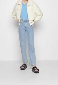 Person som bär en krämfärgad sherpa-jacka, blå t-shirt, ljusblå high-waist jeans och marina plattformssandaletter med spännen.