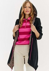 Žena v černé bundě s kapucí a podšívkou v leopardoím vzoru, oblečená v růžovém pruhovaném tričku se zipem a béžových kalhotách, držící bundu otevřenou.