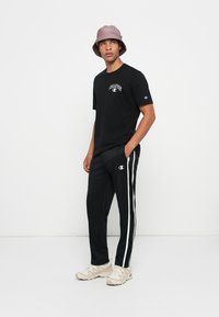 Ung mann iført en mauve bøttehatt, svart Champion t-skjorte, svarte treningsbukser med hvite striper og beige sneakers som står mot en hvit bakgrunn.