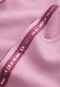 Tela rosa con un tejido texturizado. Una cinta de color púrpura oscuro presenta las palabras "BAKER by TED BAKER" en letras doradas a lo largo del borde.
