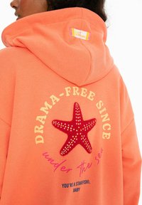 Korálová mikina s chlpatou červenou hviezdnicou. Text znie "BEZ DRÁM OD" a "pod morom," s ďalšími hravými frázami nižšie.