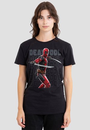 Young woman wearing black t-shirt with a colored Deadpool figure holding two swords and repeated "Deadpool" text in the background.