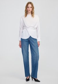 Vrouw met golvend blond haar die een witte blouse met lange mouwen, een blauwe rechte jeans en zwarte puntige hakken draagt, staande voor een effen achtergrond.