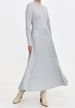 Woman wearing a light gray, long-sleeve, pleated midi dress with beige ankle boots against a plain white background.