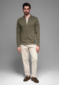 Giacca zip-up verde oliva con polsini a costine, abbinata a pantaloni color crema e scarpe in suede marroni. Presenta un logo discreto sulla manica.