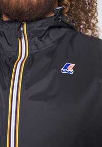 Gros plan d'une personne portant une veste noire K-Way avec une fermeture éclair rayée multicolore et le logo K-Way sur la poitrine.