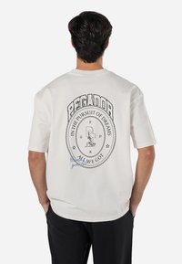White short-sleeve t-shirt featuring a bold black graphic design on the back, including the text "PEGADOR" and "IN THE PURSUIT OF DREAMS."