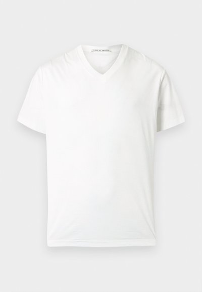 Απλή λευκή κοντομάνικη μπλούζα V-λαιμόκοψη, τοποθετημένη επίπεδη με ορατή ετικέτα στο εσωτερικό του γιακά σε ανοιχτό φόντο.