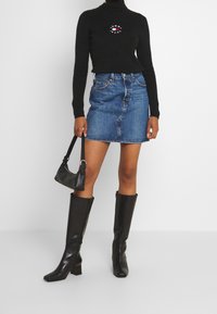 Czarny golf z detalem z logo, zestawiony z niebieską mini spódnicą z denimu i czarnymi skórzanymi kozakami sięgającymi kolan. Trzyma czarną torebkę.