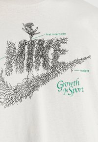 Valkoinen puuvillapaita, jossa musta juurimainen graafinen kuvio. Tekstissä on "ensimmäinen välikkö," "juuritajut" ja "Kasvu urheilussa" vihreällä.