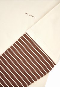 Krémová košile s dlouhými rukávy s hnědo-bílými pruhy na rukávu. Logo "OH APRIL" v hnědé blízko límce. Měkký bavlněný materiál.