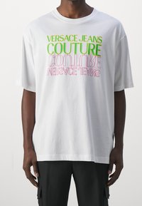 Bílý oversized tričko s barevným textem: "VERSACE JEANS COUTURE" v zelené a růžové barvě. Měkká, hladká látka s krátkými rukávy.