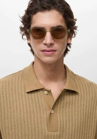 Polo marrone con un design a maglia testurizzato, due bottoni sul colletto, abbinato a occhiali da sole rotondi con una montatura chiara e lenti oscurate.