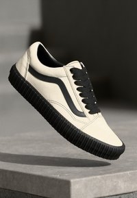 Zapatilla de deporte beige claro con acentos negros y suela de goma texturizada. Presenta cordones negros y parte superior de cuero suave con un diseño de raya lateral en contraste.