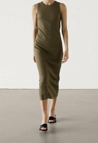 Robe midi sans manches vert olive, coupe ajustée, en tissu doux. Le modèle porte des sandales plates noires et marche sur un sol neutre.