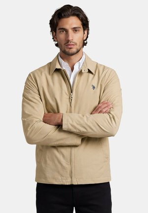 Mann mit dunklen Haaren, der eine beige Jacke mit Reißverschluss über einem weißen Hemd trägt, die Arme verschränkt, vor einem schlichten hellen Hintergrund stehend.