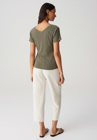 Olivgrünes Kurzarmoberteil mit V-Ausschnitt, kombiniert mit weißen Caprihosen. Modell trägt flache Sandalen und steht vor einem hellen Hintergrund.