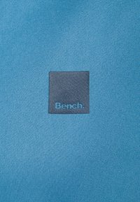 Modrá texturovaná tkanina s čtvercovou šedou záplatou, na které je dole modře vyšitý nápis „Bench.“ Jednoduchý a moderní design.