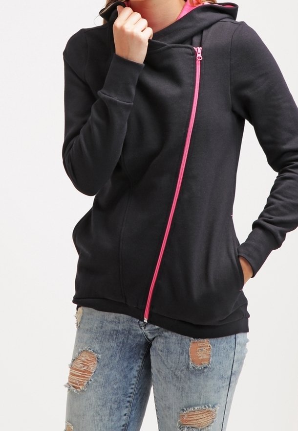 Femme portant un sweat à capuche noir avec une fermeture éclair diagonale rose et un jean bleu clair déchiré, main dans la poche et tenant le col près du visage.
