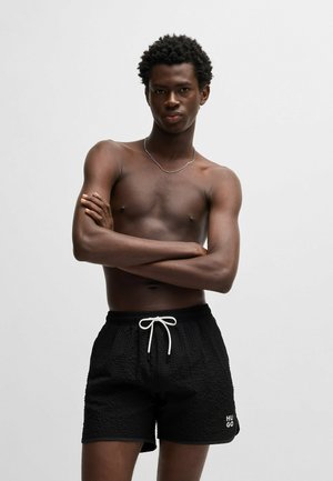Schwarze, strukturierte Badehosen mit weißem Kordelzug und Logo-Akzent. Die Shorts bieten einen lockeren Schnitt und eine kurze Länge für zusätzlichen Komfort.