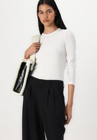 Λευκή μπλούζα με μακριά μανίκια συνδυασμένη με ψηλοκάβαλο μαύρο παντελόνι πλισέ. Το μοντέλο κρατάει μια υφασμάτινη τσάντα με μαύρη εσωτερική επένδυση και κρεμ λεπτομέρειες.
