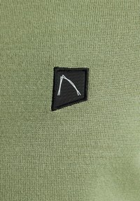 Strukturierter grüner Stoff mit einem schwarz gewebten Patch, der ein weißes geometrisches Design zeigt. Das Material ist gestrickt und erzeugt ein weiches Erscheinungsbild.