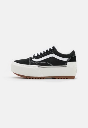 Schwarzer Sneaker mit niedrigem Schaft, weißem Wellenstreifen, dicker weißer Plateausohle, brauner profillierter Außensohle und weißen Schnürsenkeln auf einfachem, hellem Hintergrund.
