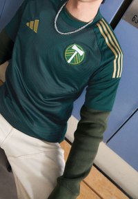 Henkilö, jolla on päällään vihreä urheilujersey, jossa on keltaisia raitoja ja logo, pitkät oliivinvihreät hihat alla, valkoiset housut ja hopeaketju.