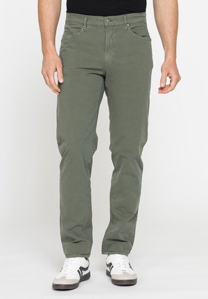 Uomo con pantaloni dritti verde oliva e sneaker bianche con dettagli neri, in piedi davanti a uno sfondo chiaro uniforme.