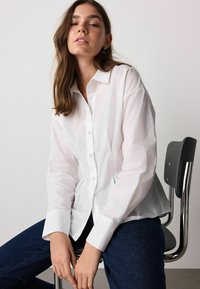White button-up shirt with long sleeves, featuring a classic collar and a straight cut. Worn with dark blue denim, sitting on a metal chair.
