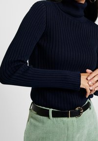 Marintröja med ribbat mönster och hög hals, långärmad, i kombination med gröna cordsbyxor, framhävt av ett svart bälte med guldspänne.