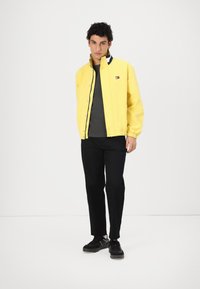 Gelbe Baumwolljacke mit Reißverschluss und Druckknopfverschluss, getragen über einem grauen Hemd. Schwarze Hosen und schwarze Sneakers runden das Outfit ab.