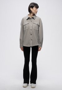 Giacca grigia con collo, chiusura a bottoni, due tasche sul petto, vestibilità comoda, indossata con pantaloni neri svasati e scarpe da ginnastica bianche.