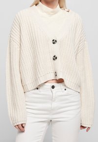 Kvinna iklädd en krämfärgad, tjockstickad, kort cardigan med stora knappar över en krämfärgad polotröja och vita högmidjade jeans.