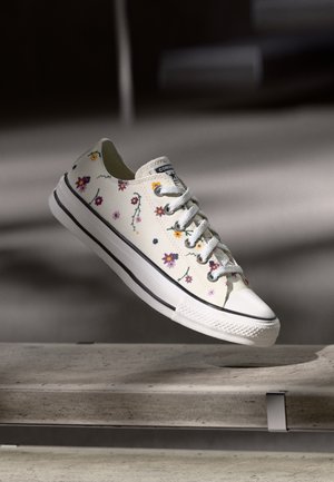Witte lage sneaker met meerkleurige bloemenprint, witte veters en een zwarte streep op de zool, zwevend boven een betonnen bank.
