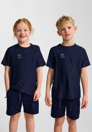 Twee kinderen, een roodharig meisje dat lacht en een blond jongetje met een neutrale gezichtsuitdrukking, beiden gekleed in bijpassende marineblauwe sportkleding met logo's op een witte achtergrond.