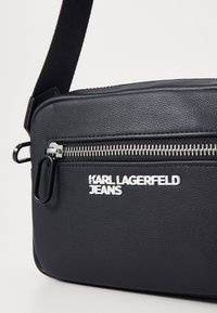 Borsa a tracolla sintetica nera con superficie texturizzata, zip in argento e logo brandizzato. Presenta una tracolla nera rimovibile.