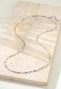 Collier en argent avec des maillons hexagonaux et ovales alternés, présenté sur une surface en pierre beige texturée avec des ombres douces.