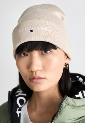 Młoda osoba w beżowej czapce z logo Tommy Jeans, złotych kolczykach-kółkach i zielonej kurtce z czarnym kapturem, patrząca prosto przed siebie.