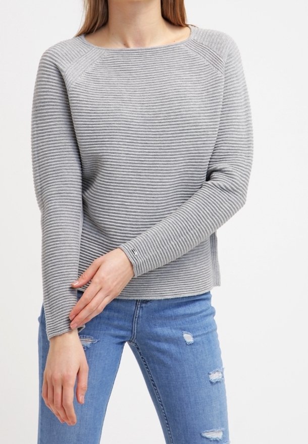 Femme portant un pull à manches longues gris côtelé et un jean bleu déchiré, ajustant la manche gauche avec sa main droite.