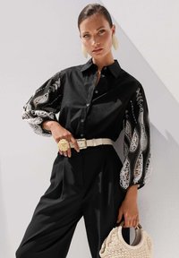 Schwarzes Hemd mit Spitzenbestickten Ärmeln, weißer Gürtel mit floralen Akzenten, schwarze Hose und eine gewebte beigefarbene Handtasche. Glatte Stoffoberfläche, figurbetontes Design.