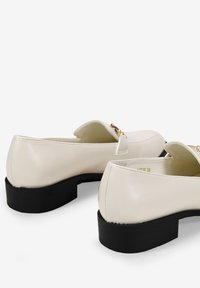 Mocasines de cuero beige con puntera redondeada, suelas de goma negra y acentos en tono dorado. Textura suave con un diseño minimalista.