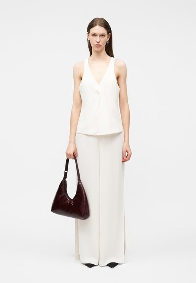 Femme portant un haut blanc sans manches à col en V et un pantalon large blanc, tenant un sac à main en cuir marron foncé, debout devant un fond uni.