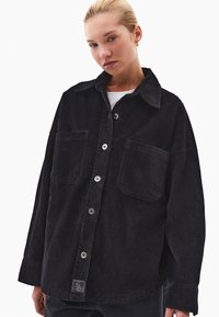 Czarna koszula z korduroy z dopasowanym kołnierzykiem, dwoma przednimi kieszeniami i zapięciem na guziki. Posiada pionową teksturę oraz metkę z logo na dolnym brzegu.