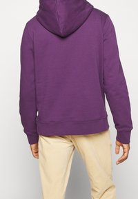 Fialová mikina s kapucí a klokaní kapsou, s manžetami a lemem v žebrovaném provedení. Bavlněný materiál s hladkou texturou, nošena s béžovými kalhotami.